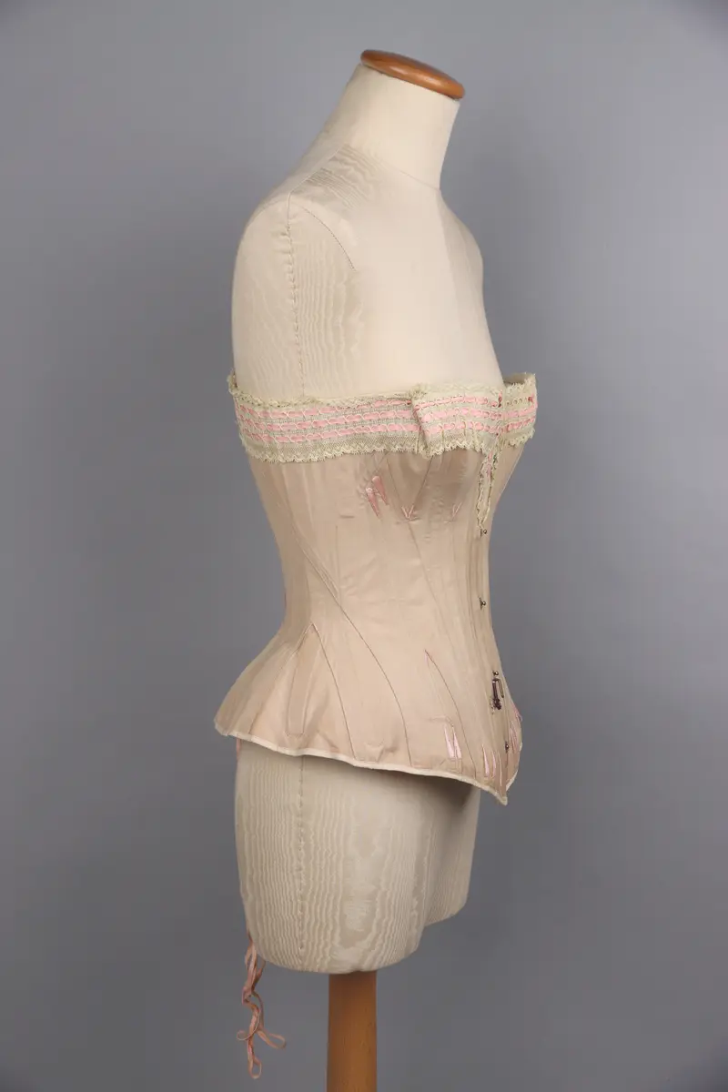 Modekorsett av begie bomull vävd i kypert. Benad med tunna fjäderstål runt hela plagget. Längs hela överkanten är en vit spets med inträdda rosa sidenband fastsydd. Svängd förnicklad planschett fram, där varje hyska är präglade med C P över "sirene". Inga övriga stämplar eller etiketter. På hela plagget är förstärkningar gjorda med rosa broderigarn, möjligen av silke. Snörning längs ryggen genom 16 par snörhål med öljetter av mässing. Snöres med platt flätad rosa snodd. På framsida och baksida finns två stora hakar fastsydda för att haka i de plagg som burits över korsetten.