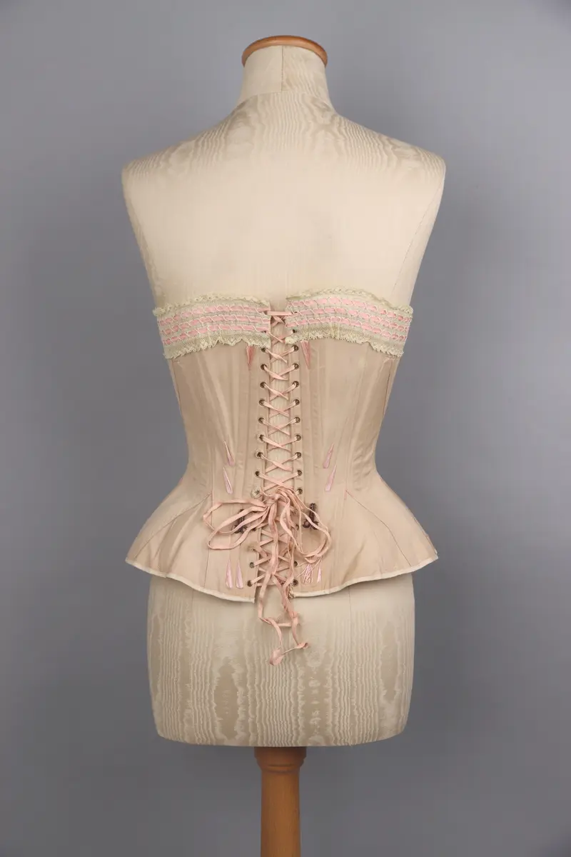 Modekorsett av begie bomull vävd i kypert. Benad med tunna fjäderstål runt hela plagget. Längs hela överkanten är en vit spets med inträdda rosa sidenband fastsydd. Svängd förnicklad planschett fram, där varje hyska är präglade med C P över "sirene". Inga övriga stämplar eller etiketter. På hela plagget är förstärkningar gjorda med rosa broderigarn, möjligen av silke. Snörning längs ryggen genom 16 par snörhål med öljetter av mässing. Snöres med platt flätad rosa snodd. På framsida och baksida finns två stora hakar fastsydda för att haka i de plagg som burits över korsetten.