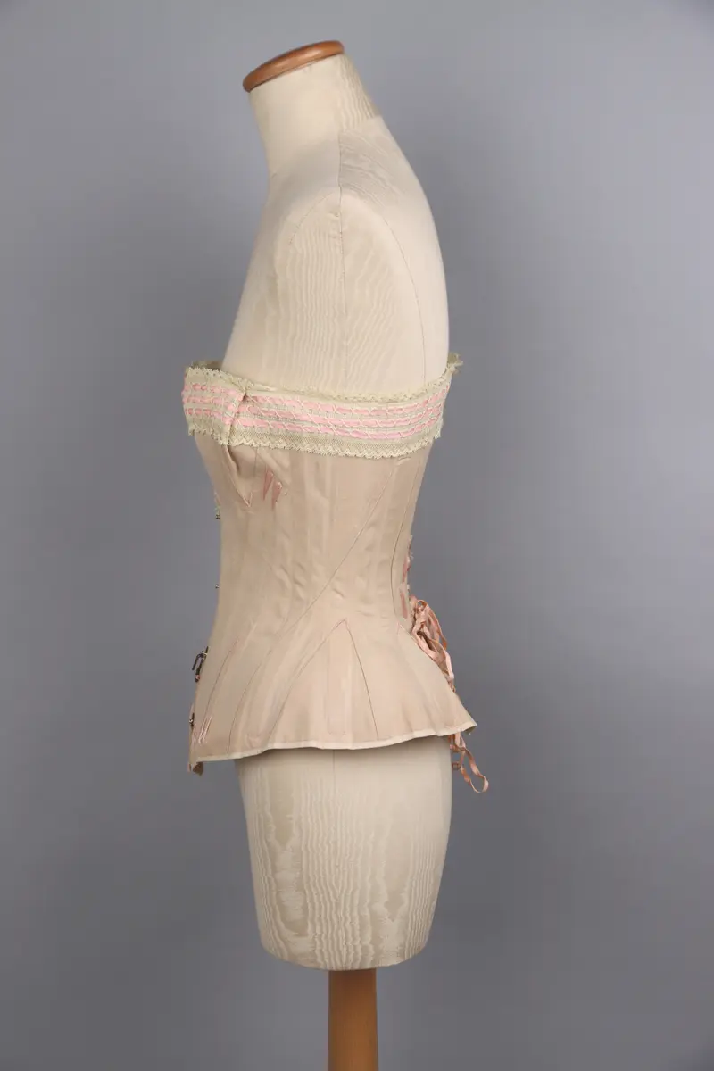 Modekorsett av begie bomull vävd i kypert. Benad med tunna fjäderstål runt hela plagget. Längs hela överkanten är en vit spets med inträdda rosa sidenband fastsydd. Svängd förnicklad planschett fram, där varje hyska är präglade med C P över "sirene". Inga övriga stämplar eller etiketter. På hela plagget är förstärkningar gjorda med rosa broderigarn, möjligen av silke. Snörning längs ryggen genom 16 par snörhål med öljetter av mässing. Snöres med platt flätad rosa snodd. På framsida och baksida finns två stora hakar fastsydda för att haka i de plagg som burits över korsetten.