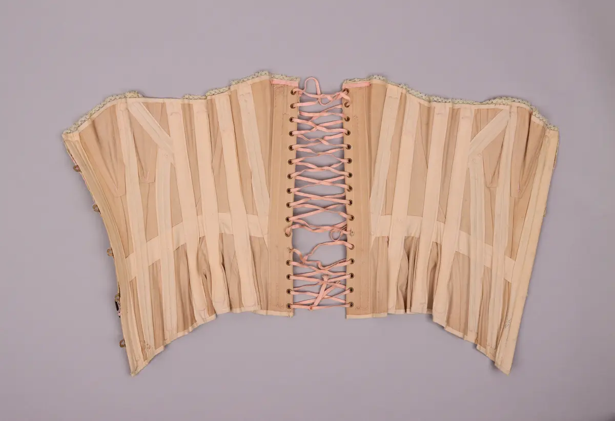 Modekorsett av begie bomull vävd i kypert. Benad med tunna fjäderstål runt hela plagget. Längs hela överkanten är en vit spets med inträdda rosa sidenband fastsydd. Svängd förnicklad planschett fram, där varje hyska är präglade med C P över "sirene". Inga övriga stämplar eller etiketter. På hela plagget är förstärkningar gjorda med rosa broderigarn, möjligen av silke. Snörning längs ryggen genom 16 par snörhål med öljetter av mässing. Snöres med platt flätad rosa snodd. På framsida och baksida finns två stora hakar fastsydda för att haka i de plagg som burits över korsetten.