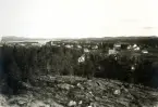 Vy över öppet landskap med bostäder, kyrka och sjö i bakgrunden samt skog och berg i förgrunden, troligen bild över Bollnäs på 1920-talet.