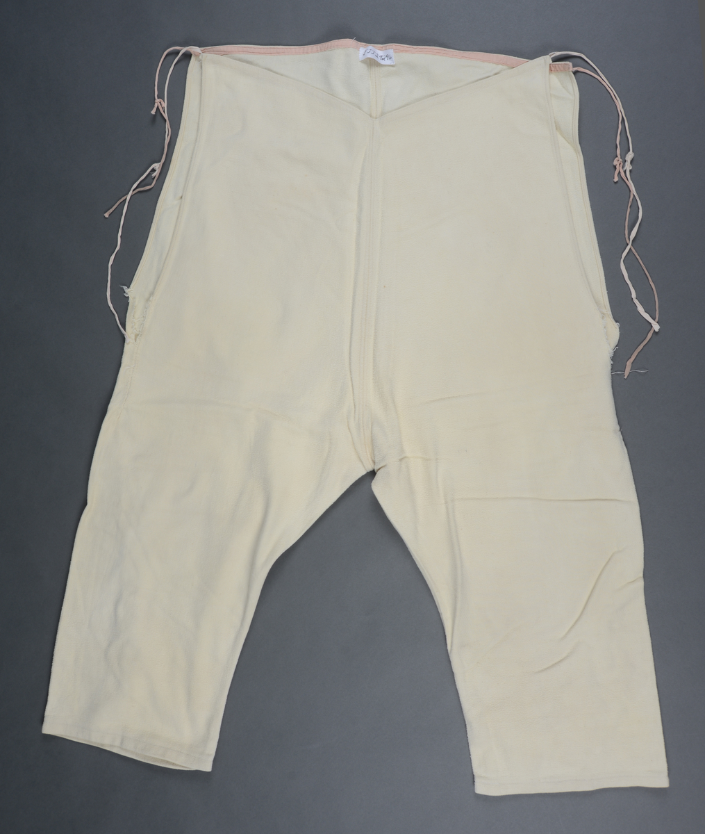 Hjemmesydd, knelang underbukse i gul/hvit flanell. Splitter i sidene, innsydd kile bakt. Bomullsbånd påsydd langs linningen, forlenget for knytting i livet. Buksa er ekstra høy i livet.