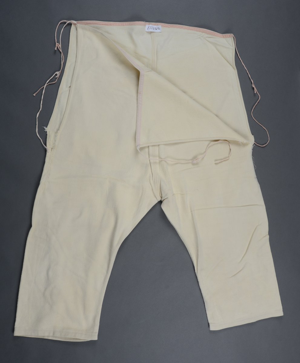 Hjemmesydd, knelang underbukse i gul/hvit flanell. Splitter i sidene, innsydd kile bakt. Bomullsbånd påsydd langs linningen, forlenget for knytting i livet. Buksa er ekstra høy i livet.