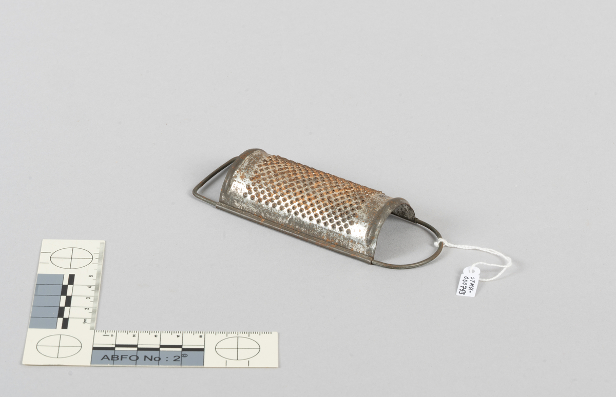 Krydderrasp. Metall. Bøyleformet. Dekket med mothakere/rasper. Gripebøyle i den ene kortenden. Plan støttebøyle i den andre.