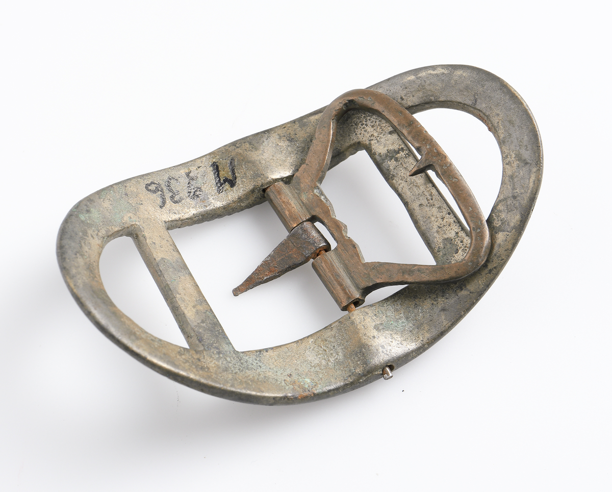 Knäspänne av järn. Oval form, något välvd. Genombruten ram i ytterkanterna, dekor på inre kant.

Inskrivet i huvudbok 1886.