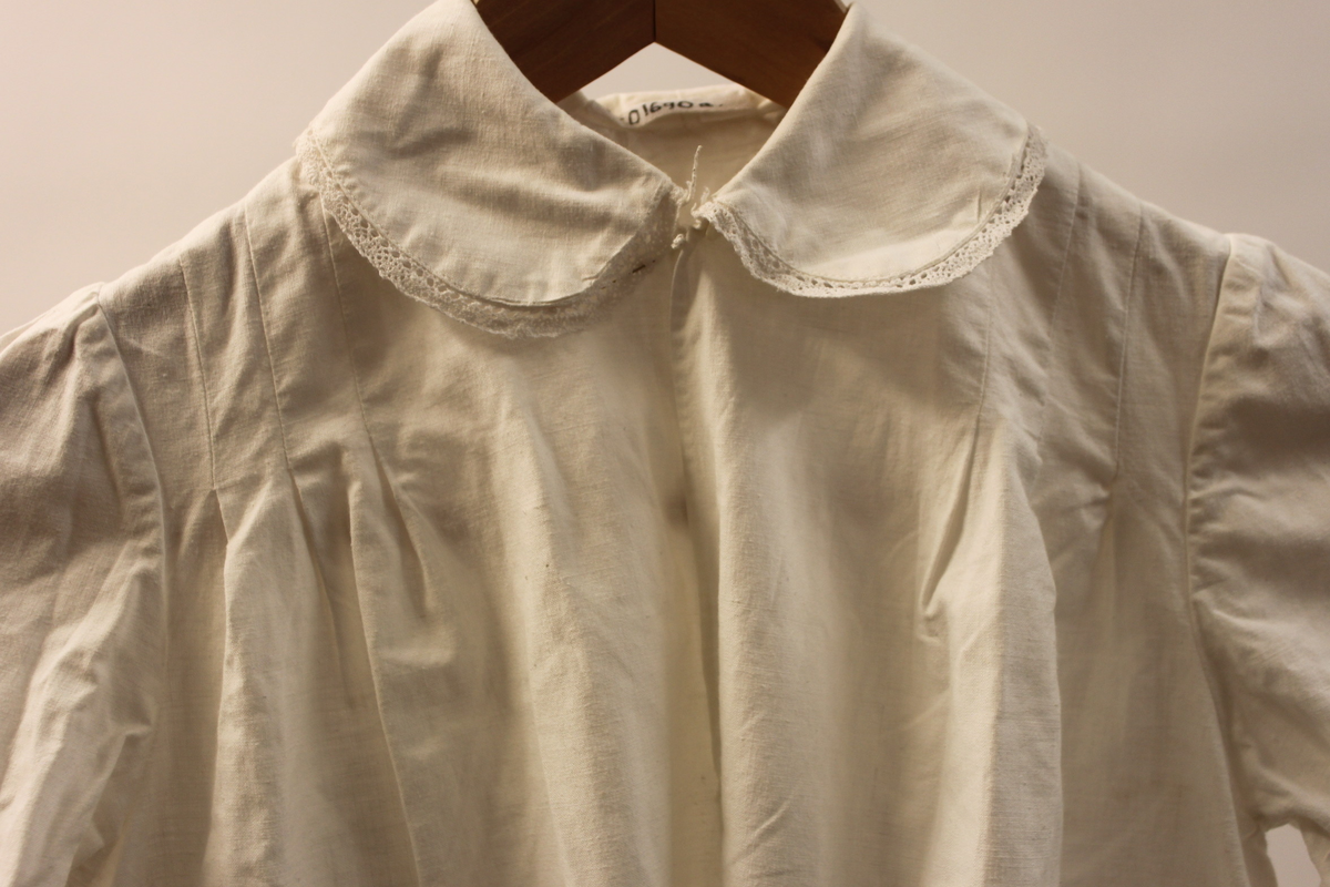 Lucialinne i bomullstyg. Barnstorlek. Hemsytt. försedd med vit spets i ärmslut och runt kragen. Sprund framtill som knäpps med en vit knapp. Tre lagda veck på bakstycket nedanför halslinningen och framtill på båda sidor om halslinningen.