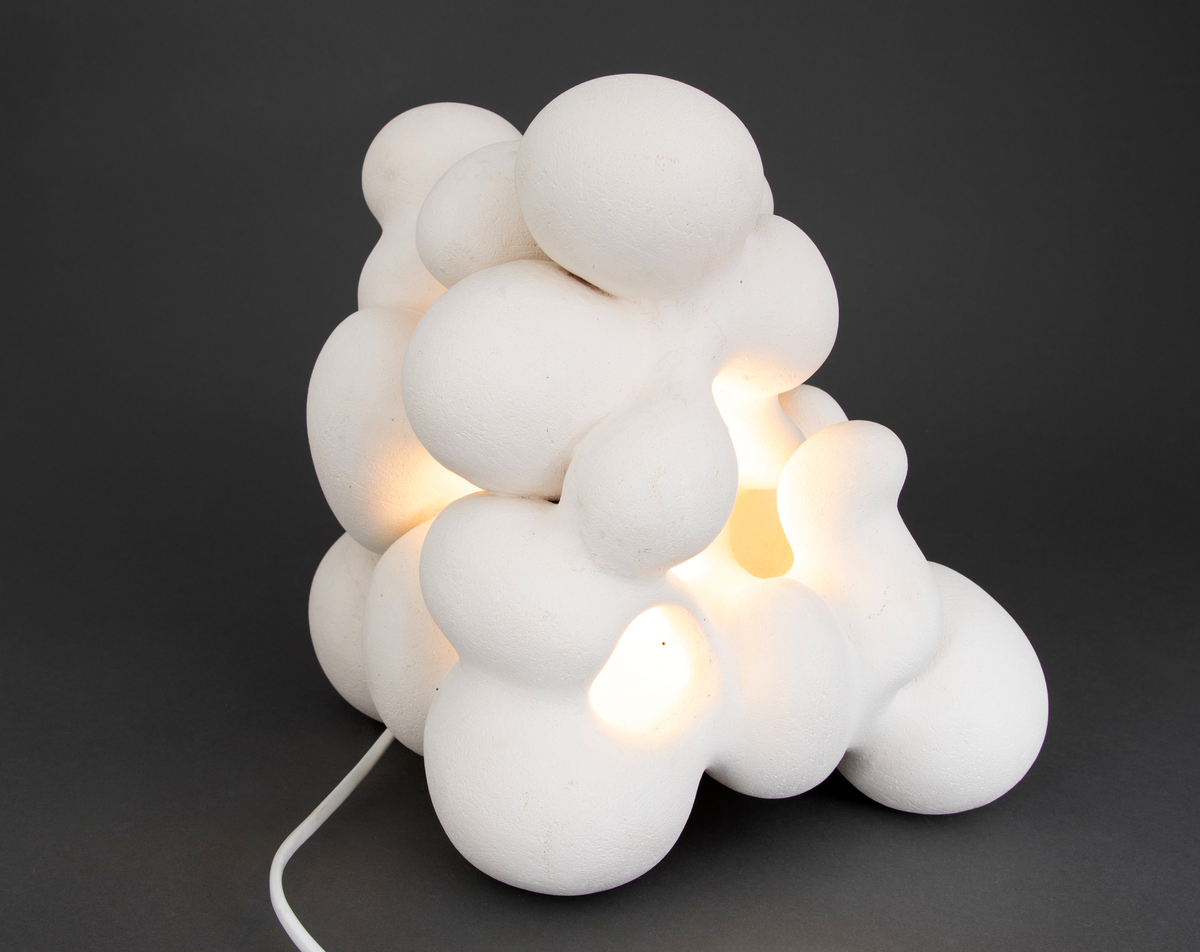 Hvit lampe i porselen. Formet av sammensmeltede bobleformer. Rund, gul lyspære inni, lyskilden er synlig fra utsiden. Skjult kabelfeste bak.