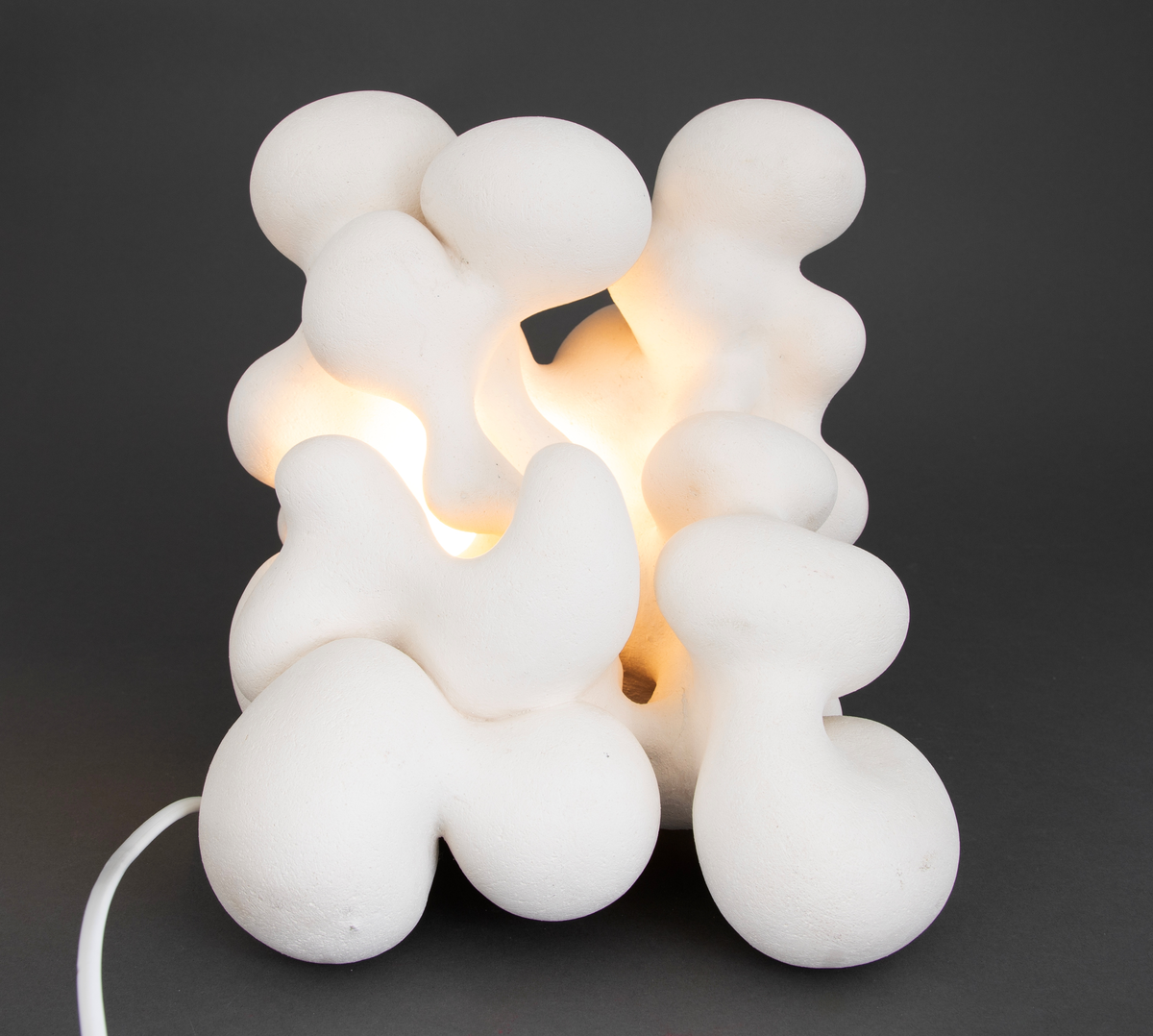 Hvit lampe i porselen. Formet av sammensmeltede bobleformer. Rund, gul lyspære inni, lyskilden er synlig fra utsiden. Skjult kabelfeste bak.