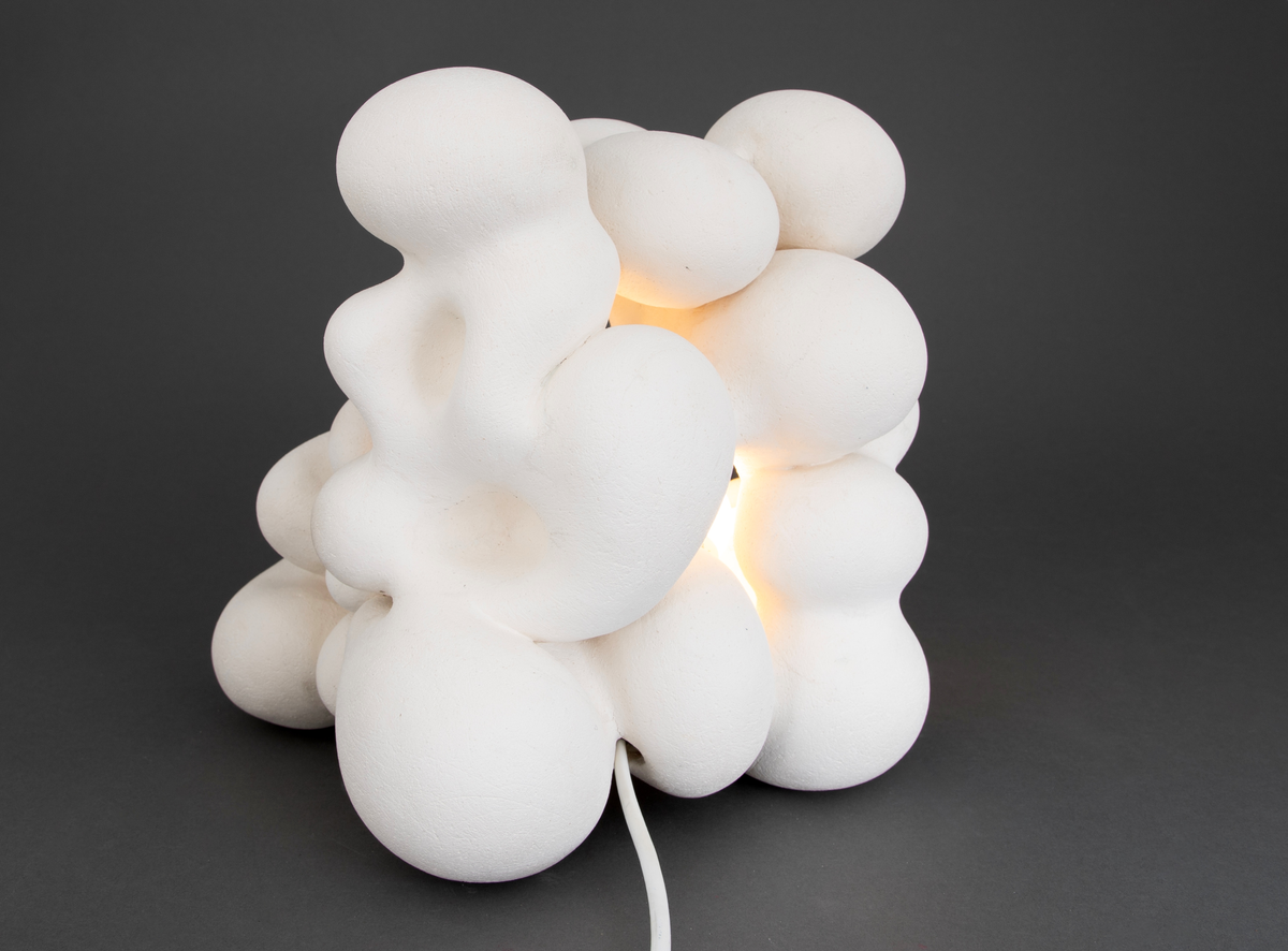 Hvit lampe i porselen. Formet av sammensmeltede bobleformer. Rund, gul lyspære inni, lyskilden er synlig fra utsiden. Skjult kabelfeste bak.