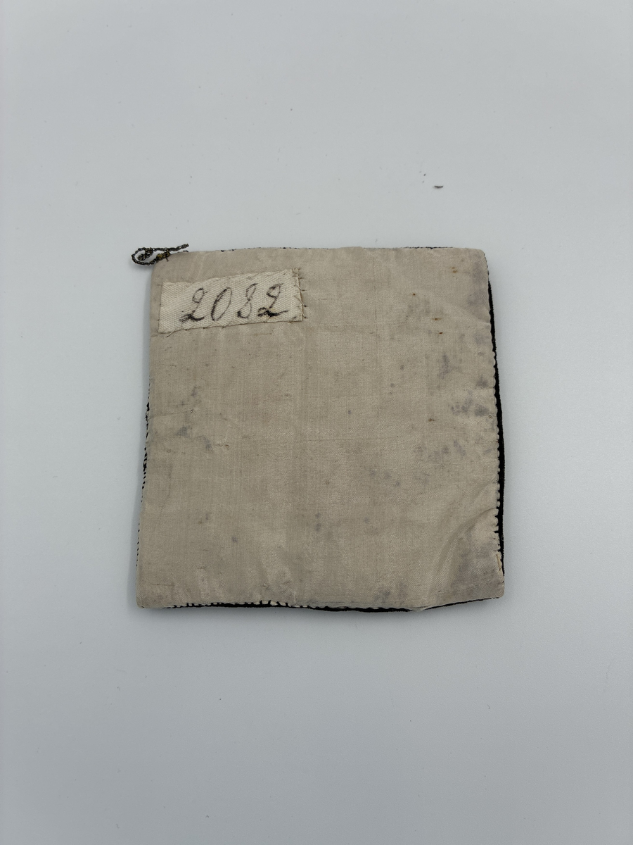 Fyrkantig dyna avsedd för placering av fickur.
Broderad och dekorerad med bl.a. fastsydda aborrfjäll.