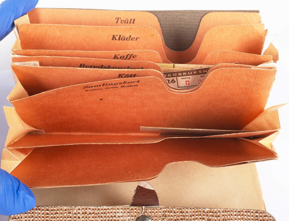 Portmonnä av papp, tryckknapp av järn och läderstropp. Använd till att förvara ransoneringskort i. Brun-gul melerad färg. Börsen har en mängd fack för de olika ransoneringskorten märkta "kaffe", "kött", "ägg", "kläder" m.m. Innehåller ransoneringskuponger av olika slag.