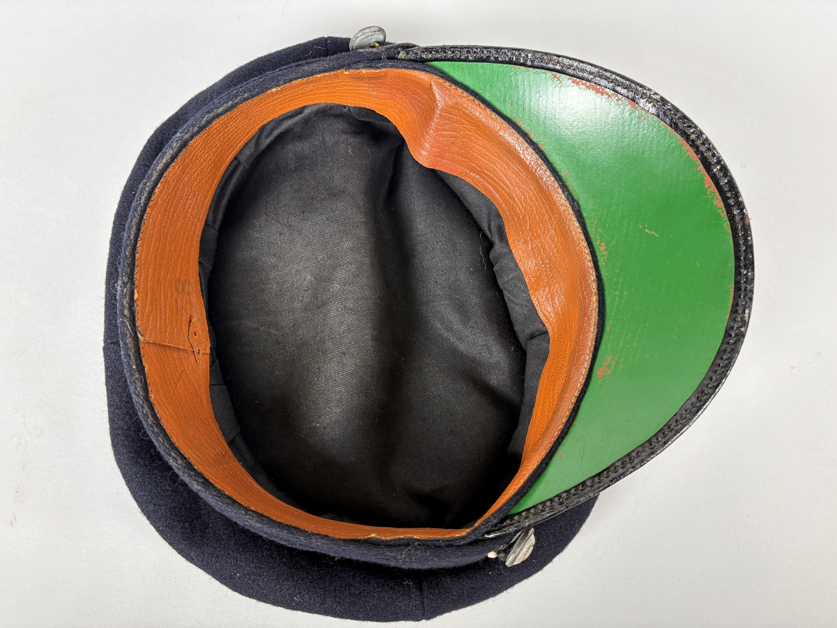 Uniformslue, mørk marineblå med sort skygge/brem. Sølvfarget bånd og Posthorn-merke i metall over skyggebrem. "Knapper" med posthorn på sidene. 

Noe bruksslitasje, mangler kokarde i nasjonalfarger.