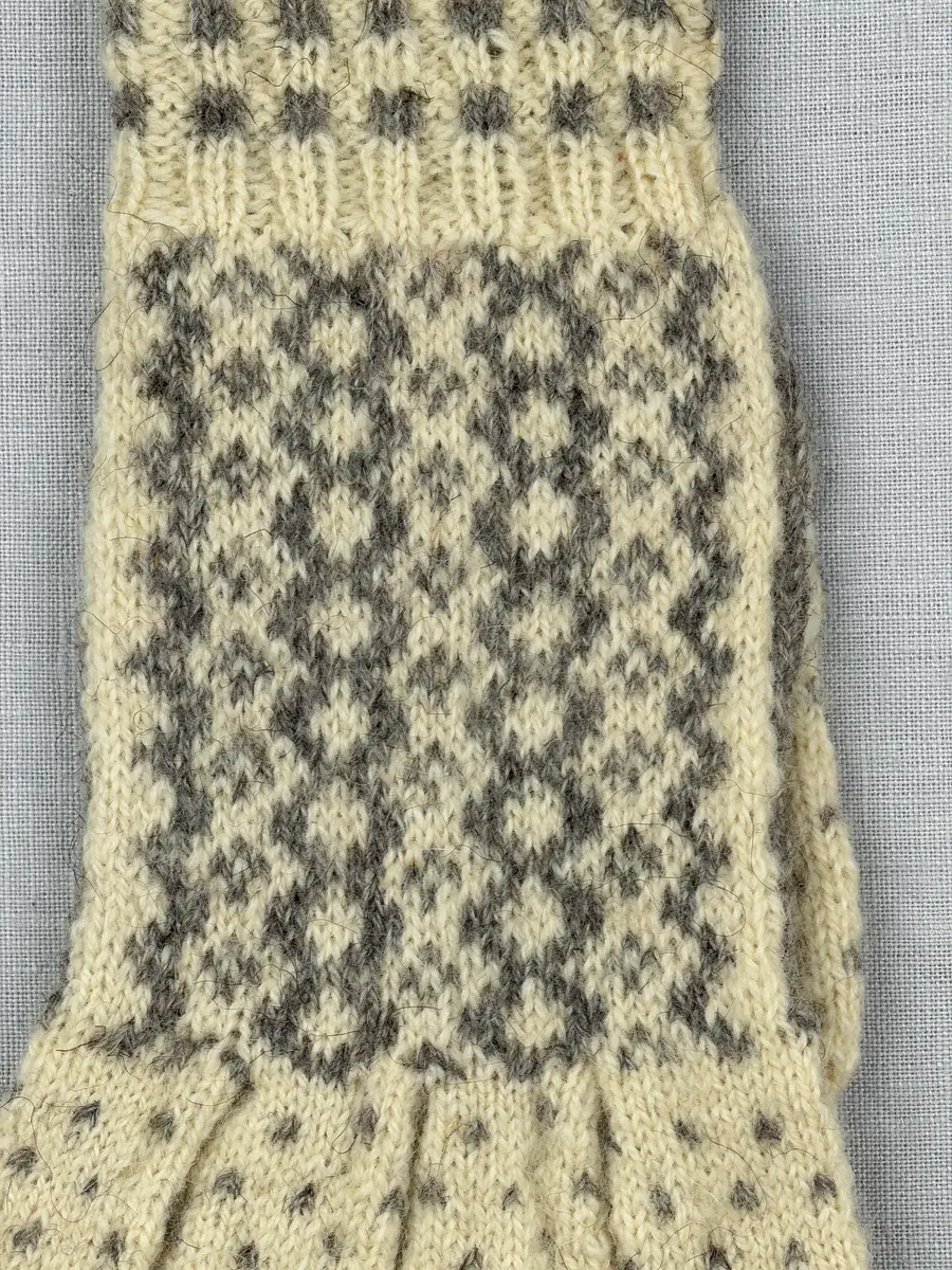 En stickad fingervante, med mönstrad mudd i 2-2 resår (2 räta och 2 aviga maskor) och mönsterstickad vante i slätstickning, av 2-trådigt ullgarn.