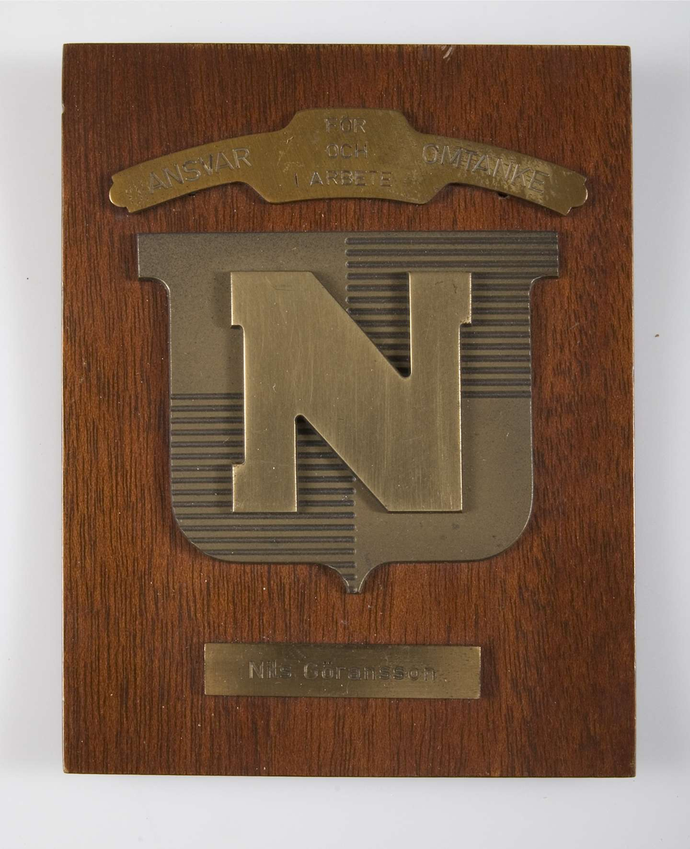 Minnesplakett med Nymanbolagens symbol, en sköld med ett N i mässing fäst på en bit ädelträ. Ovanför skölden ett band av mässing med text "För ansvar och omtanke i arbete" och under skölden en mässingsskylt med text "Nils Göransson". Upphängningskrok på baksidan och text "AB Skyltindustri, Stockholm".