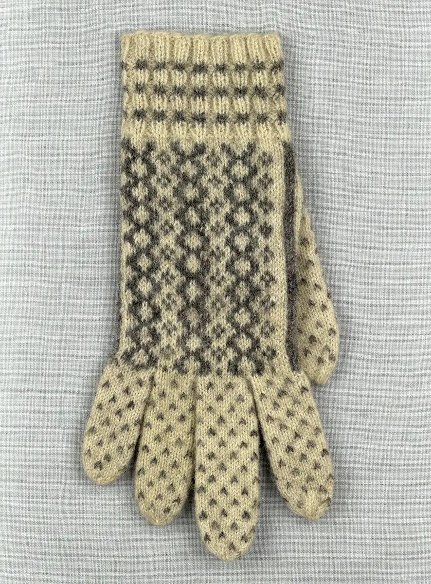 En stickad fingervante, med mönstrad mudd i 2-2 resår (2 räta och 2 aviga maskor) och mönsterstickad vante i slätstickning, av 2-trådigt ullgarn.