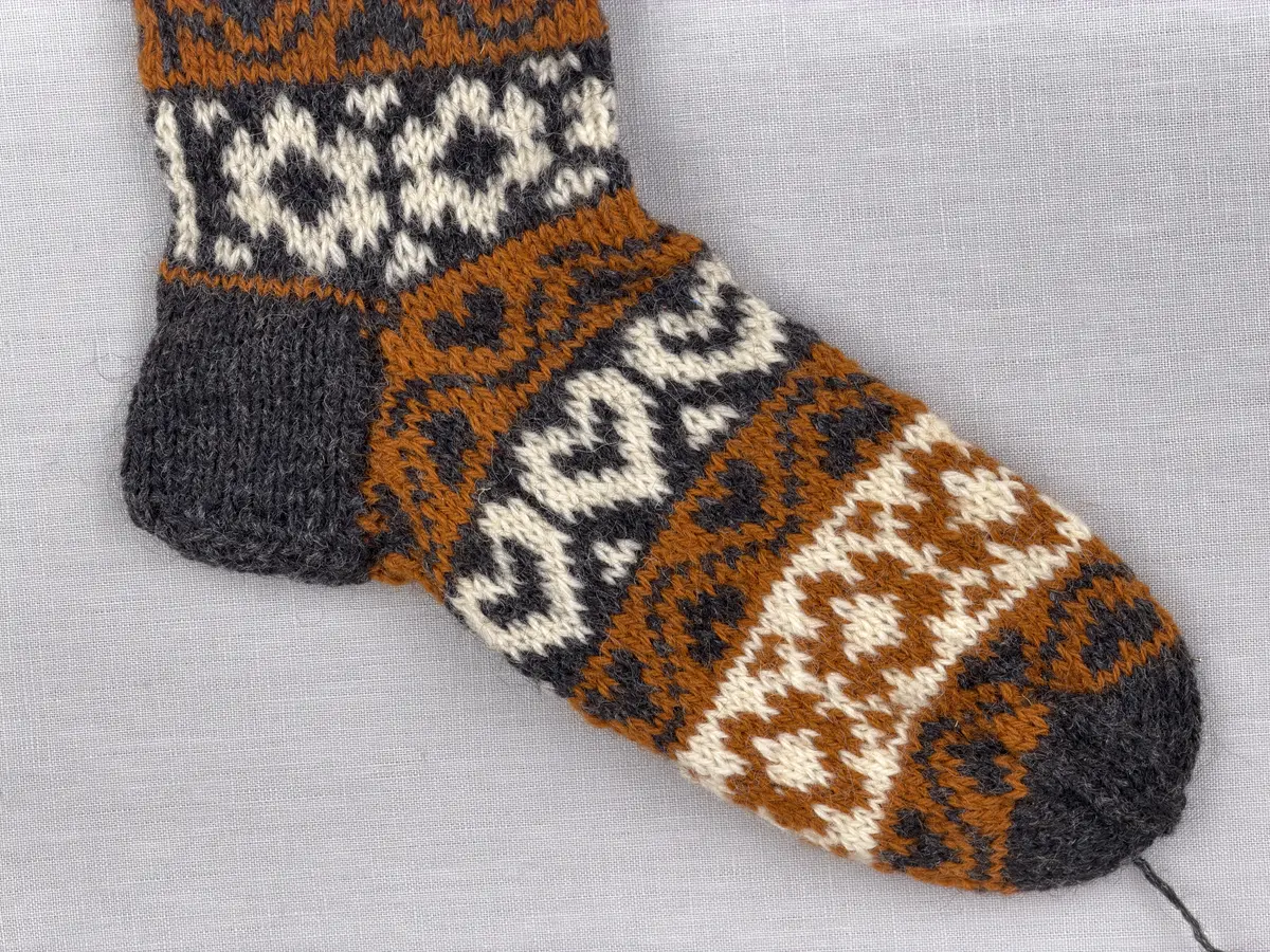 En stickad socka, med mudd i 2-2 resår (2 räta och2 aviga maskor) samt mönsterstickad fot och skaft i slätstickning, av 3-trådigt ullgarn.