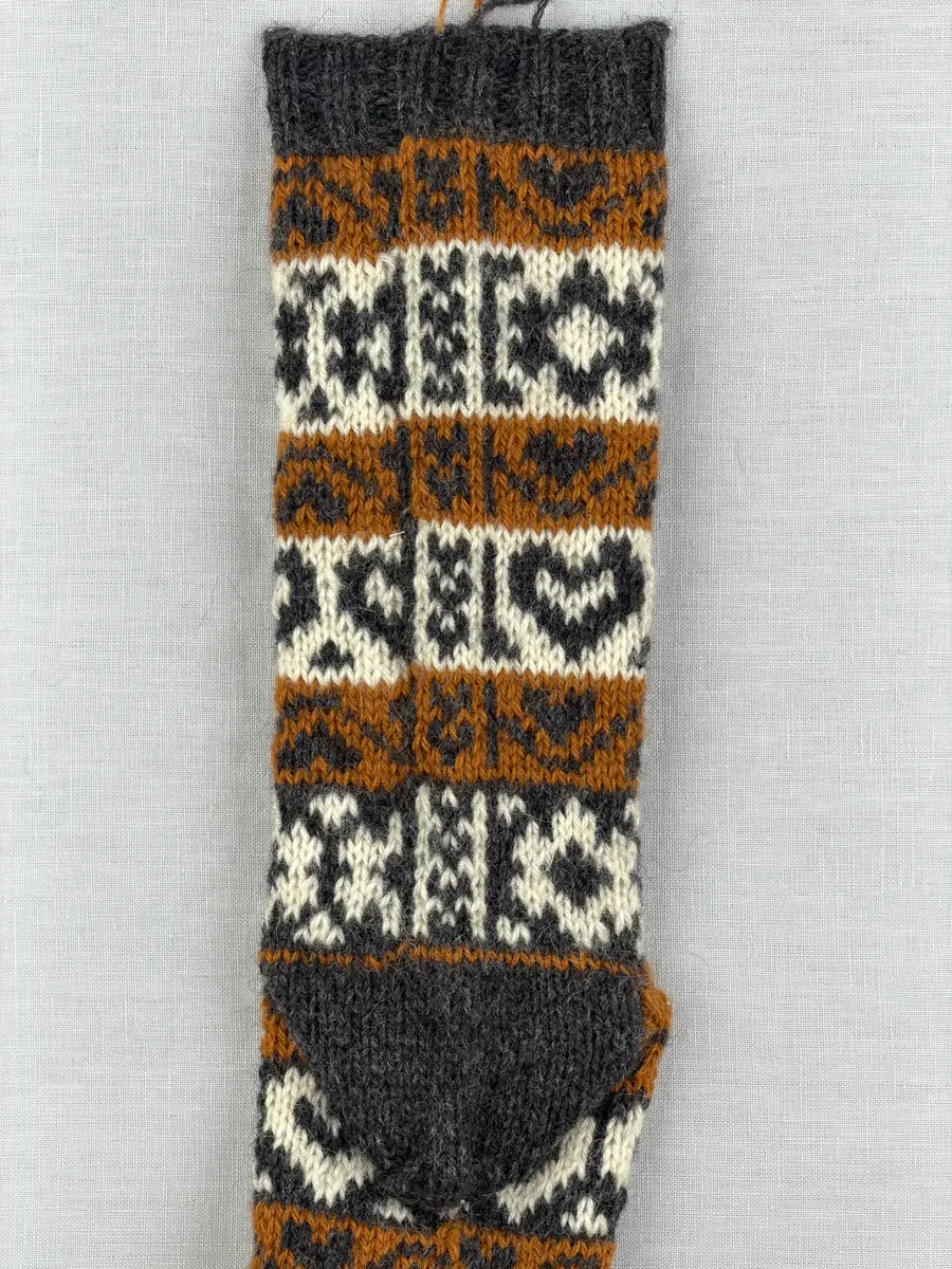 En stickad socka, med mudd i 2-2 resår (2 räta och2 aviga maskor) samt mönsterstickad fot och skaft i slätstickning, av 3-trådigt ullgarn.