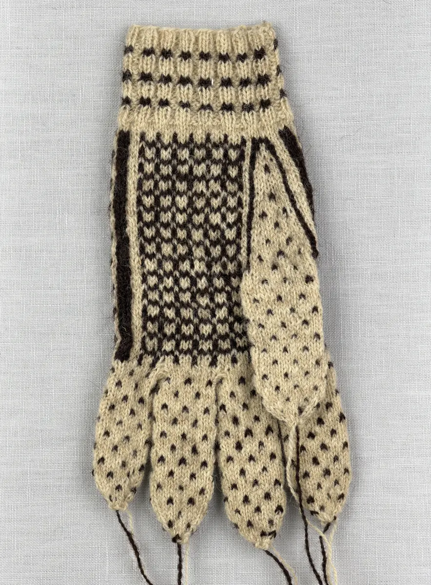 En stickad fingervante, med mönstrad mudd i 2-2 resår (2 räta och 2 aviga maskor) och mönsterstickad vante i slätstickning, av 2-trådigt ullgarn.
