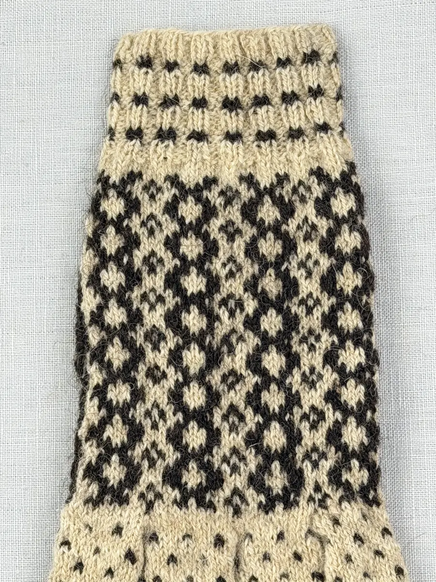 En stickad fingervante, med mönstrad mudd i 2-2 resår (2 räta och 2 aviga maskor) och mönsterstickad vante i slätstickning, av 2-trådigt ullgarn.