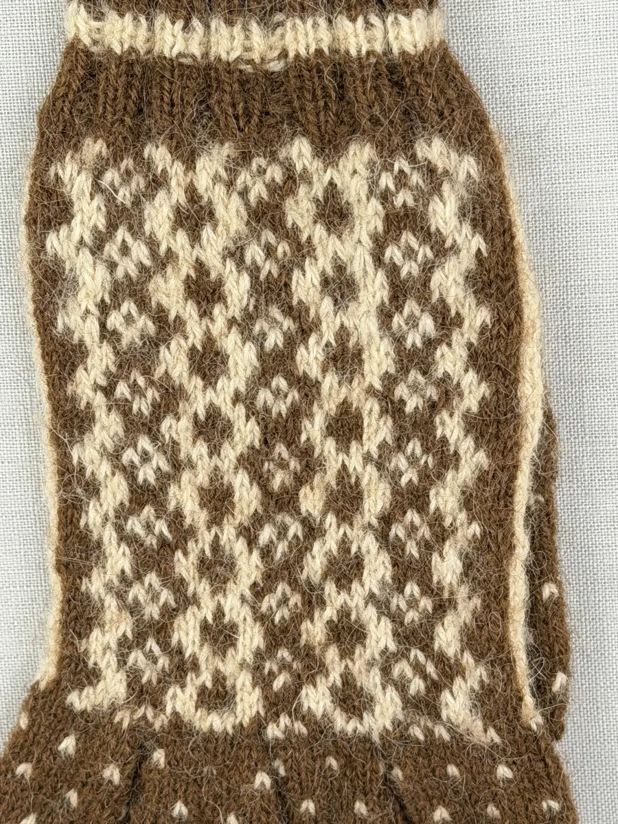 En stickad fingervante, med mönstrad mudd i 2-2 resår (2 räta och 2 aviga maskor) och mönsterstickad vante i slätstickning, av 2-trådigt ullgarn.