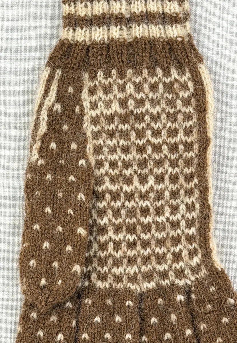 En stickad fingervante, med mönstrad mudd i 2-2 resår (2 räta och 2 aviga maskor) och mönsterstickad vante i slätstickning, av 2-trådigt ullgarn.