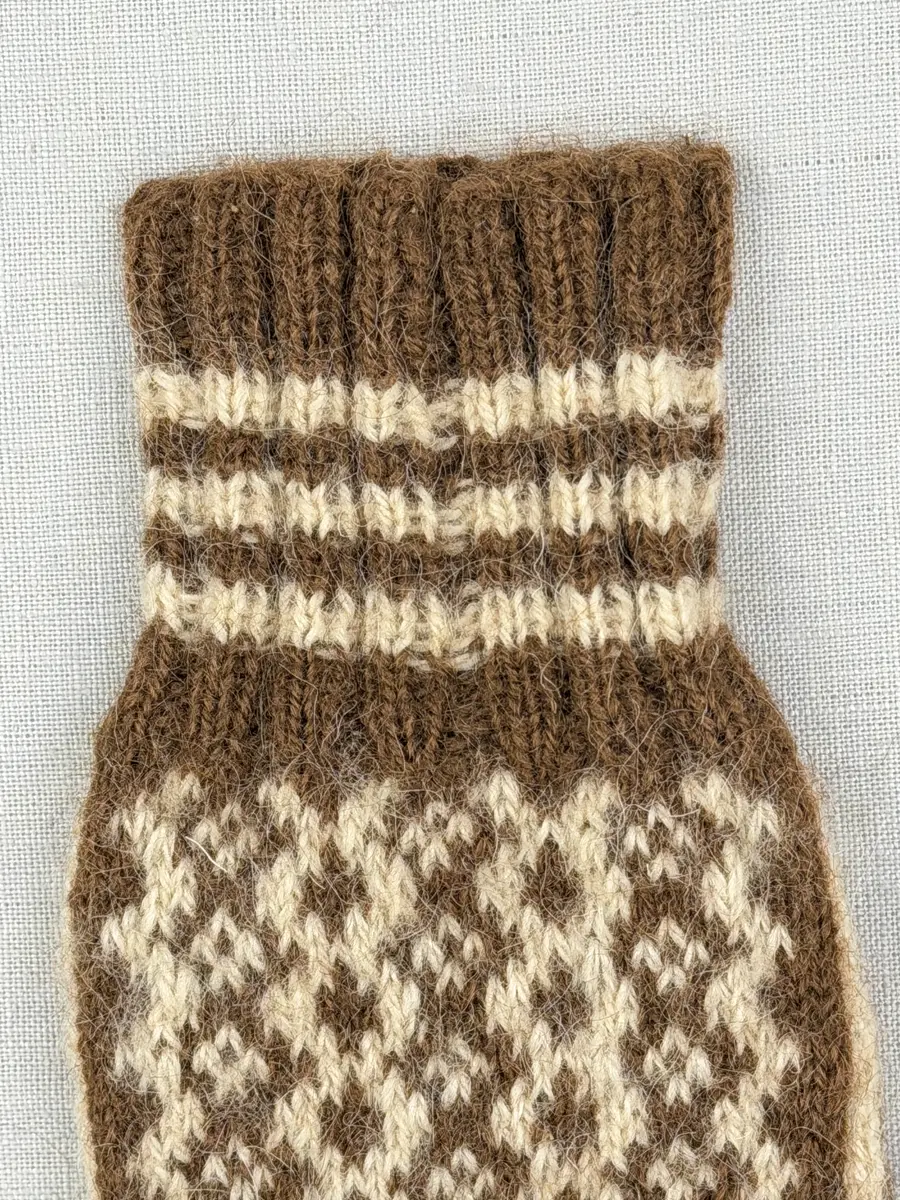 En stickad fingervante, med mönstrad mudd i 2-2 resår (2 räta och 2 aviga maskor) och mönsterstickad vante i slätstickning, av 2-trådigt ullgarn.