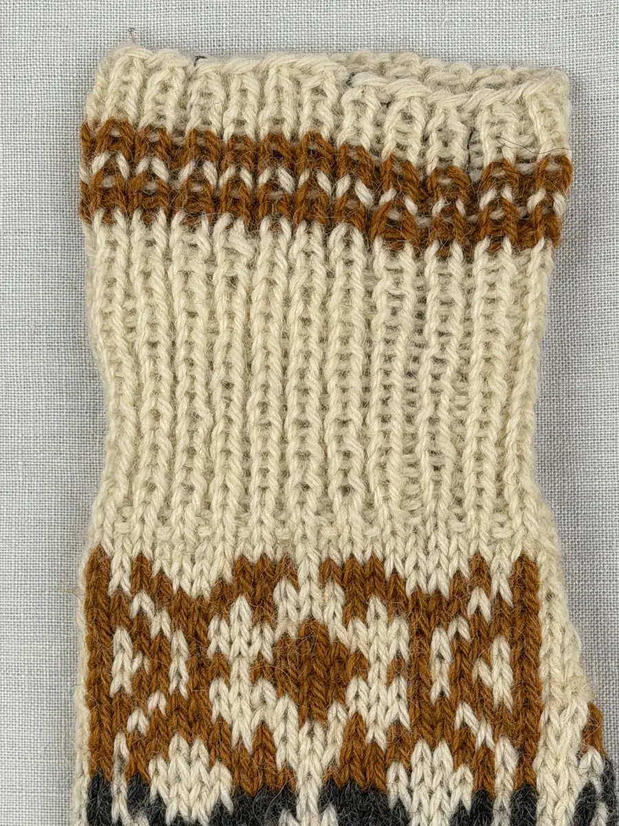 En stickad tumvante, med randig mudd i 1-1 resår (1 rät och 1 avig maska) och mönsterstickad vante i slätstickning, av 3-trådigt ullgarn.