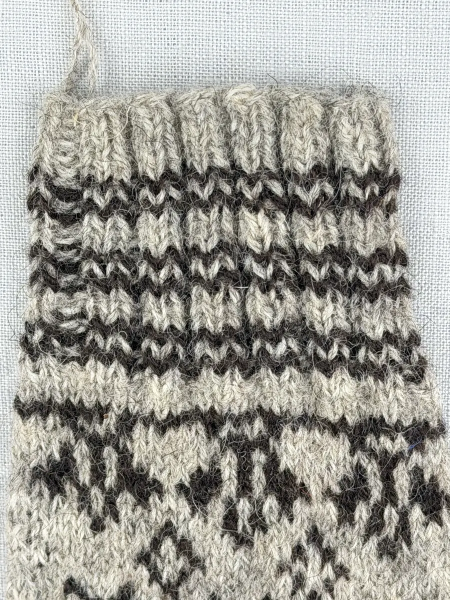 En stickad fingervante, med randig mudd i 2-2 resår (2 räta och 2 aviga maskor) och mönsterstickad vante i slätstickning, av 2-trådigt ullgarn.