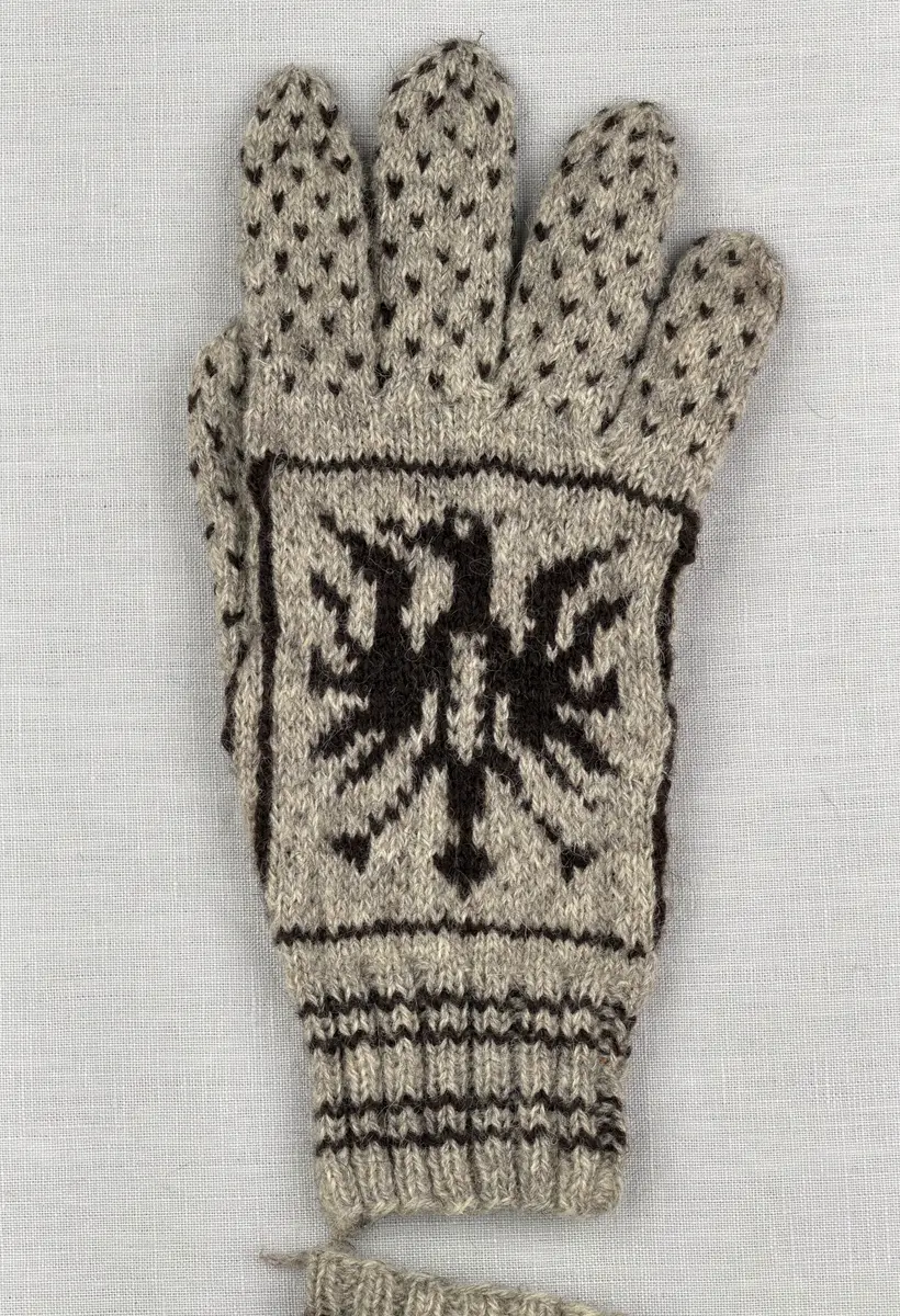 En stickad fingervante, med randig mudd i 2-2 resår (2 räta och 2 aviga maskor) och mönsterstickad vante i slätstickning, av 2-trådigt ullgarn. Framsidan är dekorerad med ett motiv föreställande Värmlandsgripen.