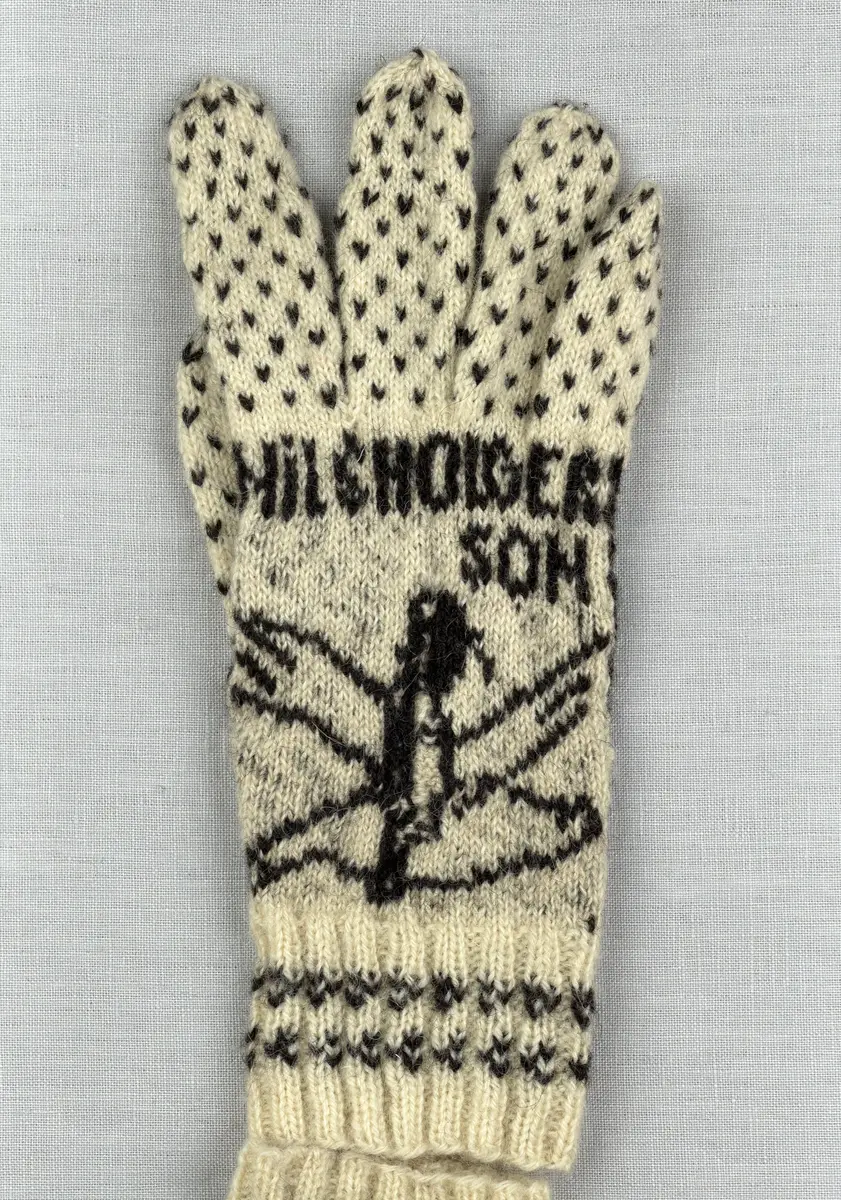 En stickad fingervante, med mönstrad mudd i 2-2 resår (2 räta och 2 aviga maskor) och mönsterstickad vante i slätstickning och aviga maskor, av 2-trådigt ullgarn. Framsidan är dekorerad med ett motiv föreställande Nils Holgerson sittande på en flygande gås.