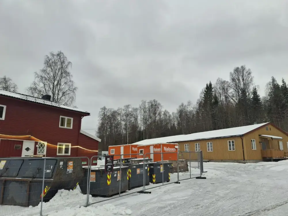 Foto av et hus med gjerde og containere foran, den gule museumsbrakka i bakgrunnen