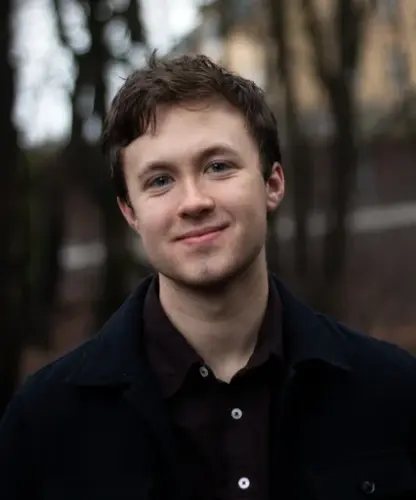 Åsmund Farstad i mørk skjorte står ute med nokre tre og eit gult bygg i bakgrunnen