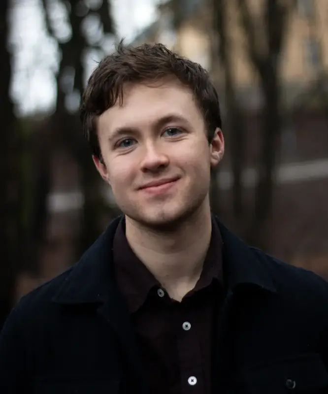 Åsmund Farstad i mørk skjorte står ute med nokre tre og eit gult bygg i bakgrunnen