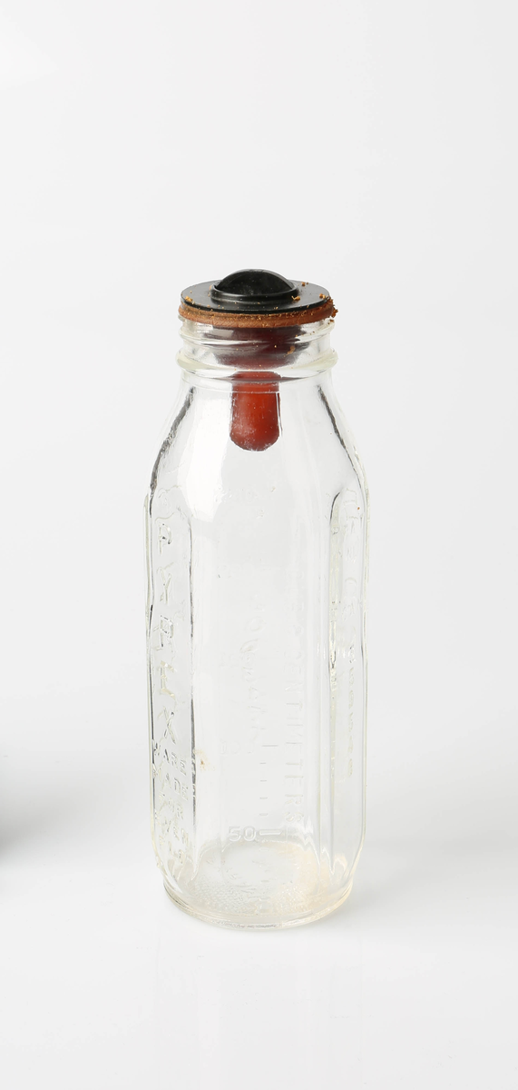 Sekssidet tåteflaske i glass med gummismokk og plastkork. Flasken er av gjennomsiktig glass, korken sort og smokken rødbrun.