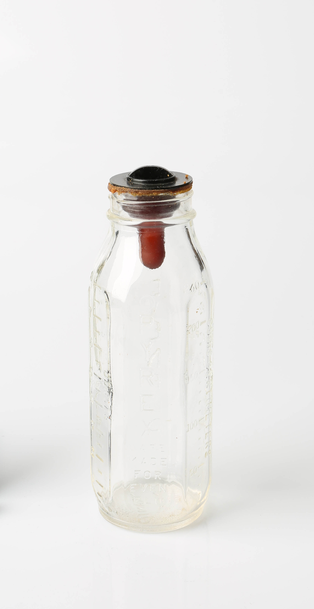 Sekssidet tåteflaske i glass med gummismokk og plastkork. Flasken er av gjennomsiktig glass, korken sort og smokken rødbrun.
