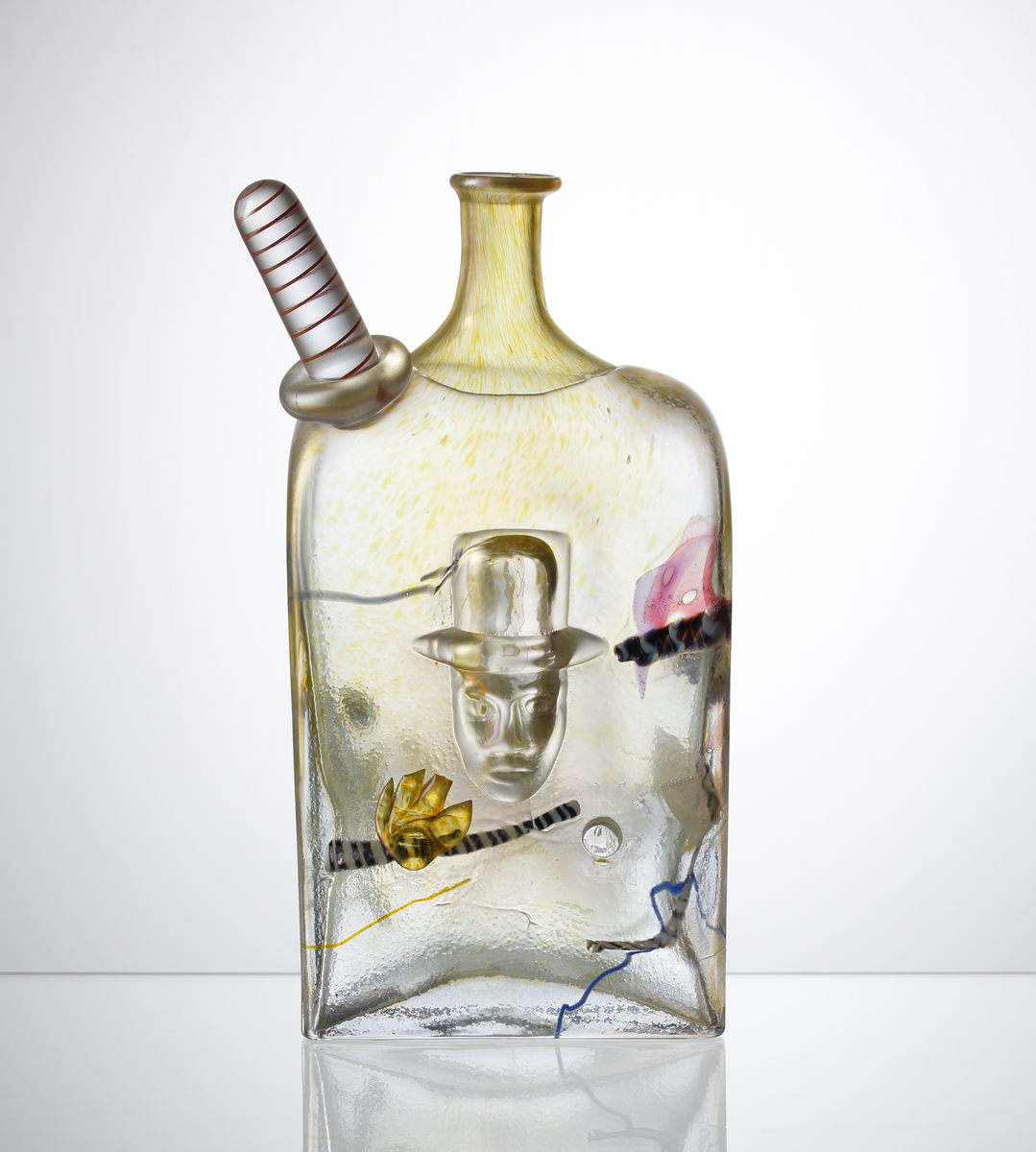 Flaska, fyrkantig.
Brunskimrande med inslag av lila.
Hamrad yta p g a uppblåst i järnform.
Ansikte i mitten, påklippt rödvit figur på skuldran och invälsade 
geometriska figurer i flaskan.
Driven.
Funktion: Prydnadsföremål