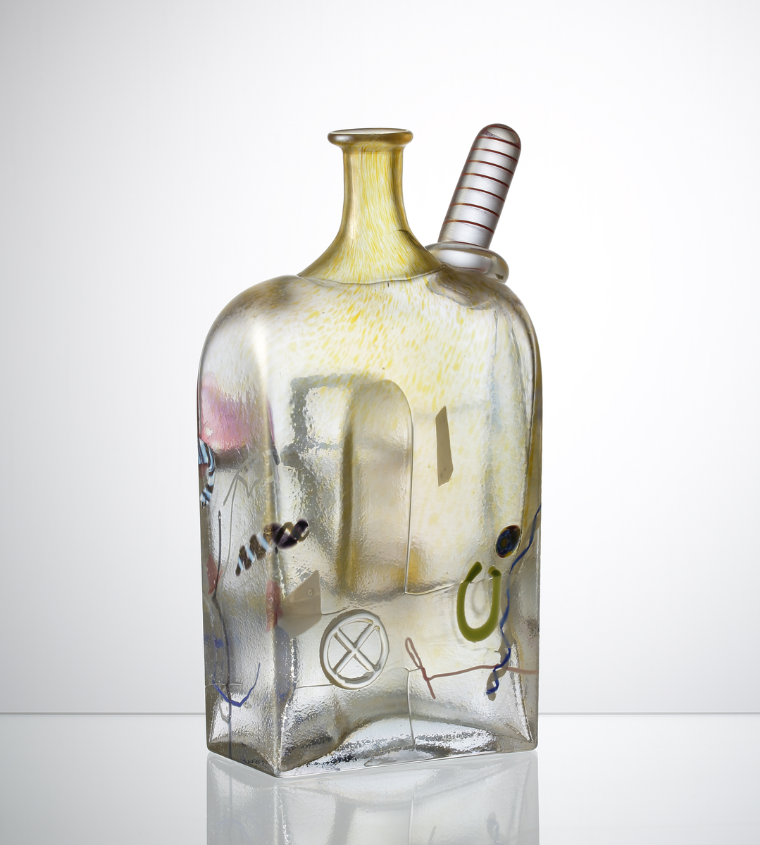 Flaska, fyrkantig.
Brunskimrande med inslag av lila.
Hamrad yta p g a uppblåst i järnform.
Ansikte i mitten, påklippt rödvit figur på skuldran och invälsade 
geometriska figurer i flaskan.
Driven.
Funktion: Prydnadsföremål