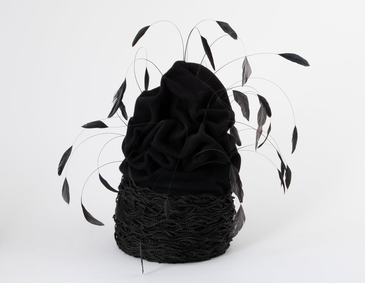 Svart hatt av turbanform, formet med dype folder og bulker. Produsert av ullfilt, med dekor av flettet nylon og vaiende, stripsete hanefjær. De smale flettene er drapert rundt hattens nedre parti, som bølgende, tettstilte "rastafletter". Hatten er meget avstivet, dvs at filten er innsatt/tilført en stivelse og er ganske grunn innvendig. Øvre del er fylt med hardt materiale. Hatten er fôret med svart silke og nederkanten påsydd et 2,5 cm bredt svettebånd, et såkalt ripsbånd. Hanefjærene er gjennomsnittlig ca 20 cm lange og stripset så kun 5 cm av fjærene står igjen ytterst, som små "dusker".