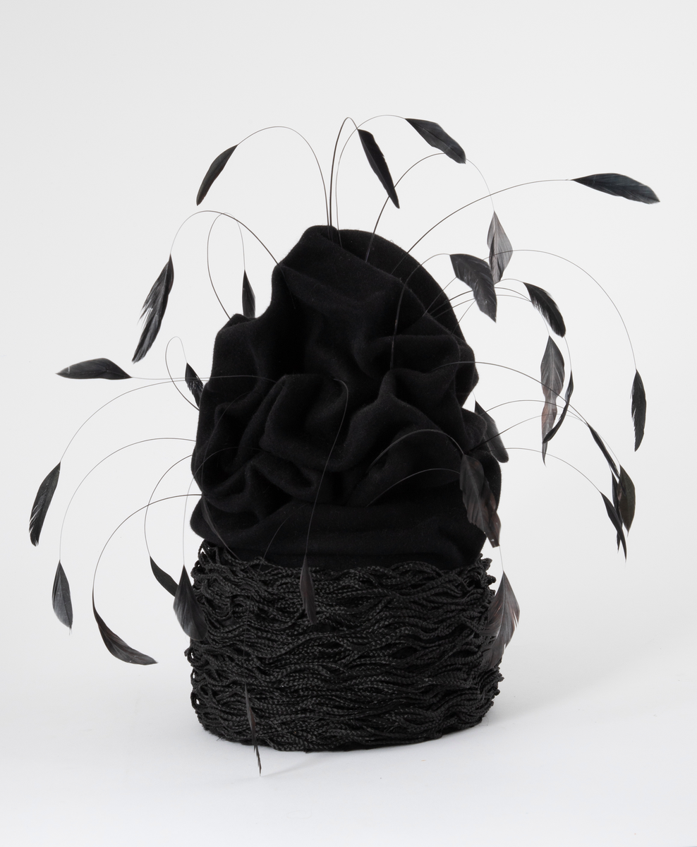 Svart hatt av turbanform, formet med dype folder og bulker. Produsert av ullfilt, med dekor av flettet nylon og vaiende, stripsete hanefjær. De smale flettene er drapert rundt hattens nedre parti, som bølgende, tettstilte "rastafletter". Hatten er meget avstivet, dvs at filten er innsatt/tilført en stivelse og er ganske grunn innvendig. Øvre del er fylt med hardt materiale. Hatten er fôret med svart silke og nederkanten påsydd et 2,5 cm bredt svettebånd, et såkalt ripsbånd. Hanefjærene er gjennomsnittlig ca 20 cm lange og stripset så kun 5 cm av fjærene står igjen ytterst, som små "dusker".
