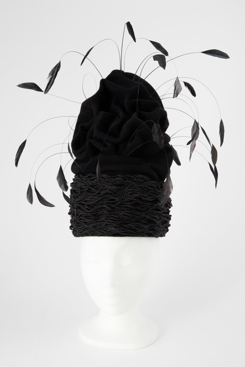 Svart hatt av turbanform, formet med dype folder og bulker. Produsert av ullfilt, med dekor av flettet nylon og vaiende, stripsete hanefjær. De smale flettene er drapert rundt hattens nedre parti, som bølgende, tettstilte "rastafletter". Hatten er meget avstivet, dvs at filten er innsatt/tilført en stivelse og er ganske grunn innvendig. Øvre del er fylt med hardt materiale. Hatten er fôret med svart silke og nederkanten påsydd et 2,5 cm bredt svettebånd, et såkalt ripsbånd. Hanefjærene er gjennomsnittlig ca 20 cm lange og stripset så kun 5 cm av fjærene står igjen ytterst, som små "dusker".