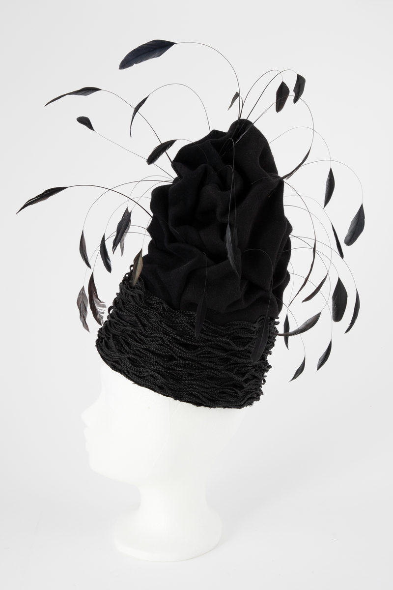 Svart hatt av turbanform, formet med dype folder og bulker. Produsert av ullfilt, med dekor av flettet nylon og vaiende, stripsete hanefjær. De smale flettene er drapert rundt hattens nedre parti, som bølgende, tettstilte "rastafletter". Hatten er meget avstivet, dvs at filten er innsatt/tilført en stivelse og er ganske grunn innvendig. Øvre del er fylt med hardt materiale. Hatten er fôret med svart silke og nederkanten påsydd et 2,5 cm bredt svettebånd, et såkalt ripsbånd. Hanefjærene er gjennomsnittlig ca 20 cm lange og stripset så kun 5 cm av fjærene står igjen ytterst, som små "dusker".