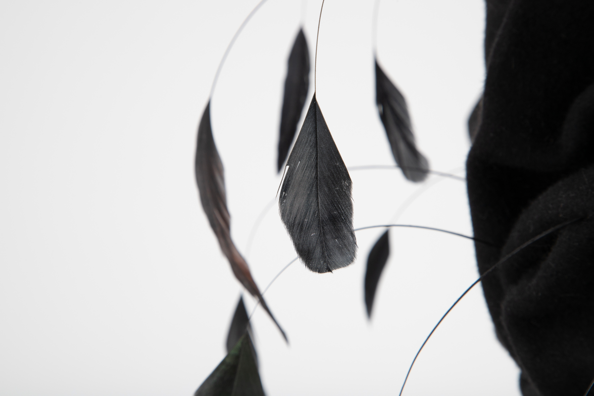 Svart hatt av turbanform, formet med dype folder og bulker. Produsert av ullfilt, med dekor av flettet nylon og vaiende, stripsete hanefjær. De smale flettene er drapert rundt hattens nedre parti, som bølgende, tettstilte "rastafletter". Hatten er meget avstivet, dvs at filten er innsatt/tilført en stivelse og er ganske grunn innvendig. Øvre del er fylt med hardt materiale. Hatten er fôret med svart silke og nederkanten påsydd et 2,5 cm bredt svettebånd, et såkalt ripsbånd. Hanefjærene er gjennomsnittlig ca 20 cm lange og stripset så kun 5 cm av fjærene står igjen ytterst, som små "dusker".