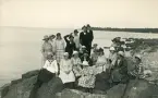 En grupp personer, både män och kvinnor, som sitter på en klippformation någonstans i någon skärgård, tyder på tidigt 1900-tal med tanke på klädseln.