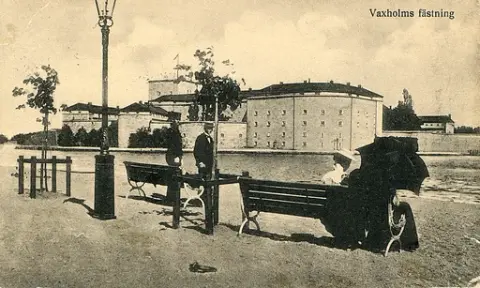 Bild på Vaxholms fästning i Stockholms skärgård med människor på promenad längs med kajen. Text baksida kort: "Vaxholms Fästning. Bästa kamrat, jag har nu kommit fram till fästningen. Det är bra kamrater jag har fått bara busiga grabbar. Piano har vi i dagrummet så där sitter vi och klinkar om dagarna. Jag skall skriva mera sedan. Min adress är (väldigt otydlig adress) Vaxholmen. Hälsa grabbarna".