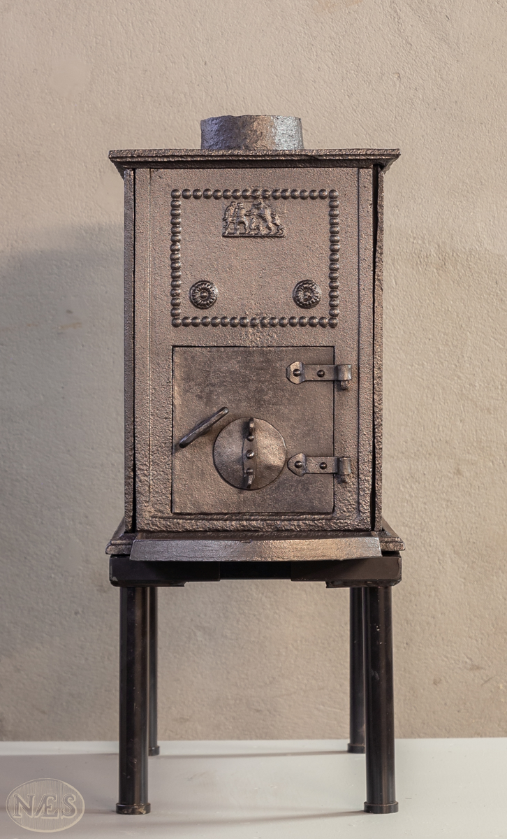 Ovn med ovnsdøren på kortsiden og røykuttak i topplaten. På langsideplatene  to amoriner i en oval krans omkranset av perler og med en krans i hvert hjørne. Ytterst en perlerand som rammer inn det hele. På Kortsiden bak er det en påskrift. Over ovnsluken på motsatt side er det et motiv med menneske og dyr innrammet i en firkantet perlerand. I de to nedre hjørnene innenfor randen er det en rosett.