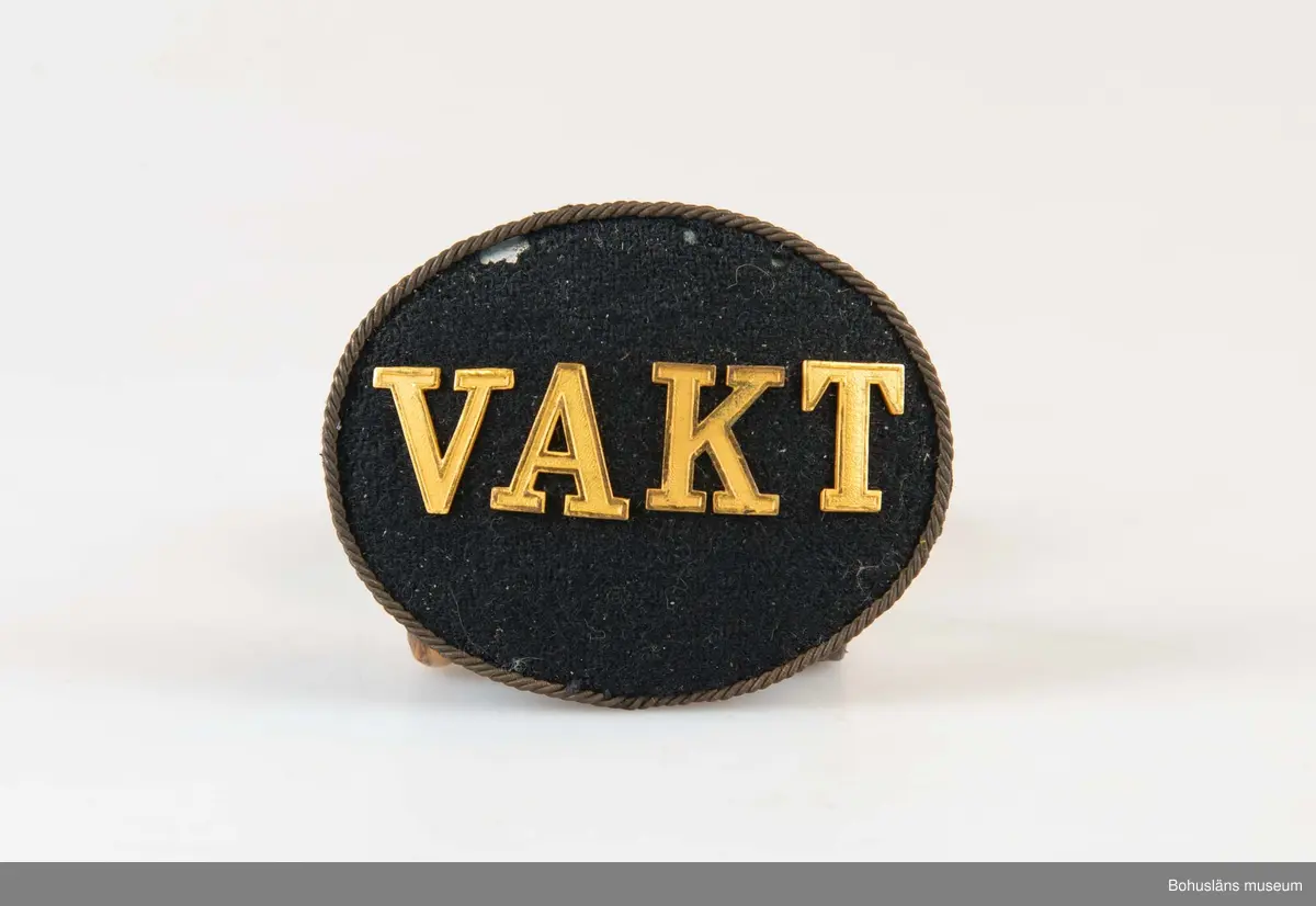Ovalt och något välvt mössmärke med texten VAKT i förgyllda mässingsbokstäver på svart sammetsbotten. band runtom. På baksidan en kraftig nål med hyska.

Märket samhör med skinnjacka och batong som använts i vakttjänstgöringen på  Uddevallavarvet.
