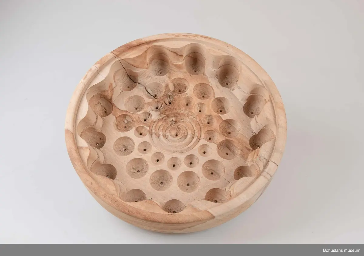 Rund, stor svarvad äggostform i lövträ med symmetriskt runda mönster och borrade hål. Utformad med hjälp av träborr av olika dimension. Linjedekor på utsidan.
På undersidan märkt med brännpenna: 
Harry Hallberg 
Ellös samt på sidan 
HH 1991.
Spricka.