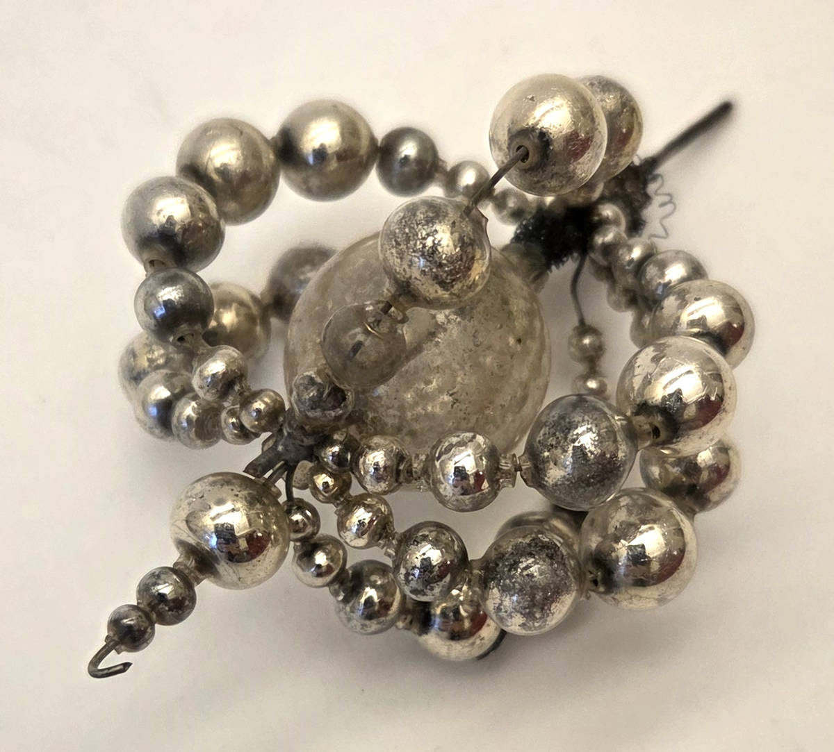 Julgranspynt i form av en glaskula omgiven av silverfärgade kulor trädda på ståltråd. En metalltråd med liten krok i ena änden löper genom glaskulan.