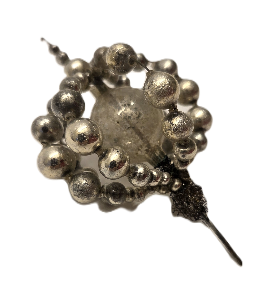 Julgranspynt i form av en glaskula omgiven av silverfärgade kulor trädda på ståltråd. En metalltråd med liten krok i ena änden löper genom glaskulan.