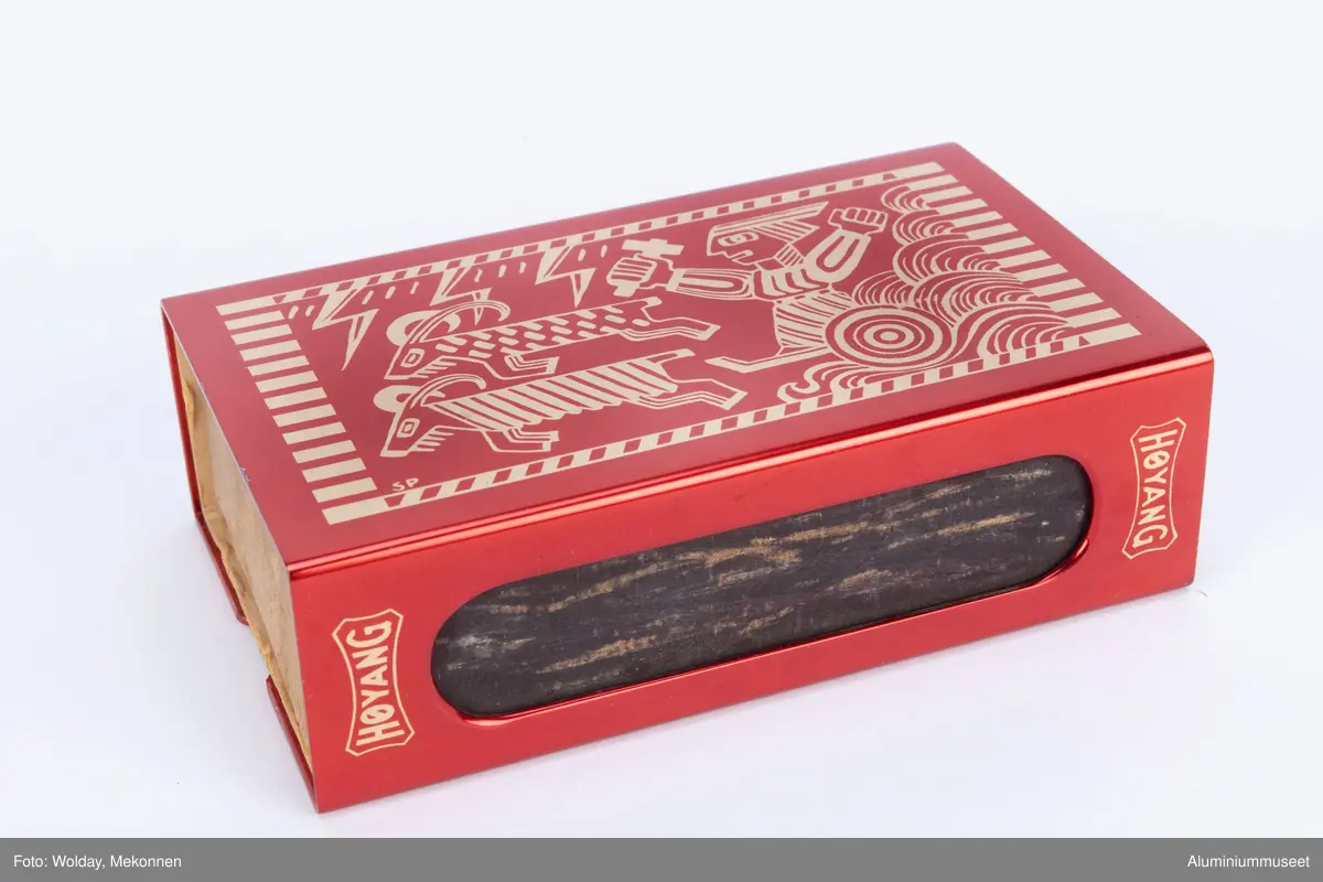 Rød, eloksert aluminium, med motiv Tor med hammeren.

Teknikk: En Aluminiumsplate ble hjørnestanset (avrundet) og to lange hull for ripeflate ble utstanset. Holderen ble knekket i to operasjoner i eksenterpresse. Først ble bunn knekket i en operasjon og så ble topp/side knekket i en operasjon. Begge operasjonene ble utført i eksenterpresse. Holderen ble polert, eloksert og farvet gul. Motivet " Tor med hammeren" ble siketrykket på med et dekkskjikt. Holderen ble så rødfarvet, men rødfarven angrep ikke motivet som var dekket. Eloksskiktet ble tilslutt tettet i sealeren (varmt vann). 
Form: Tilpasset utvendig form for stor fyrstikkeske